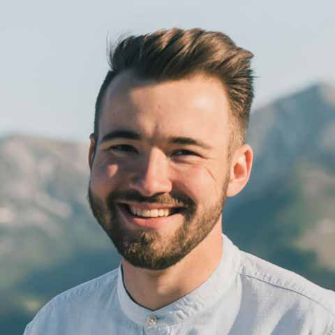 A young caucasian man with brown hair and beard smiles at the camera with mountains far away in the background
