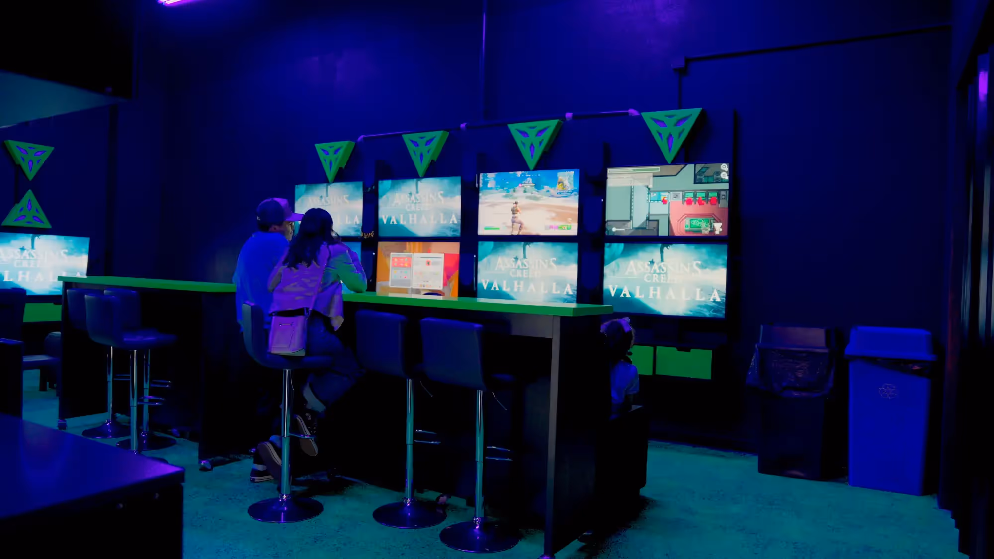 A man and woman sit at a bar with video games on the screens in front of them.