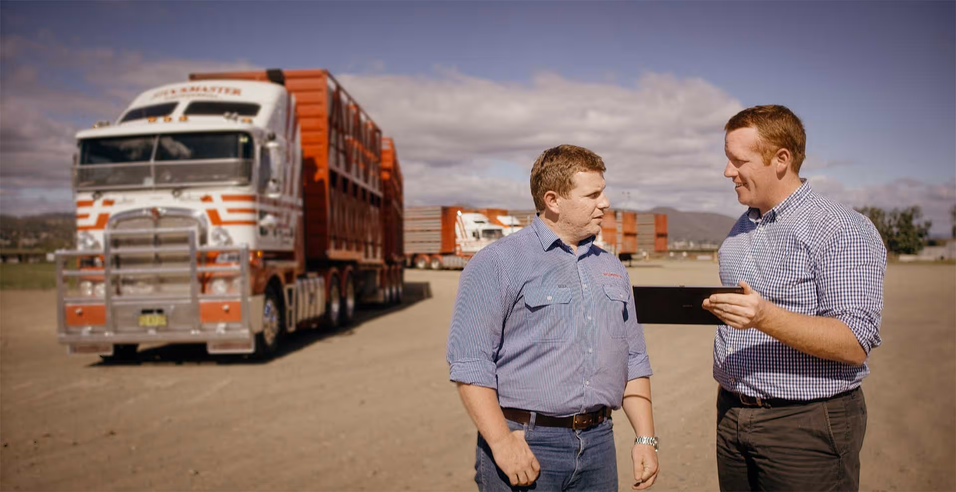 Two men in casual attire stand on a dirt road and behind them is a red truck