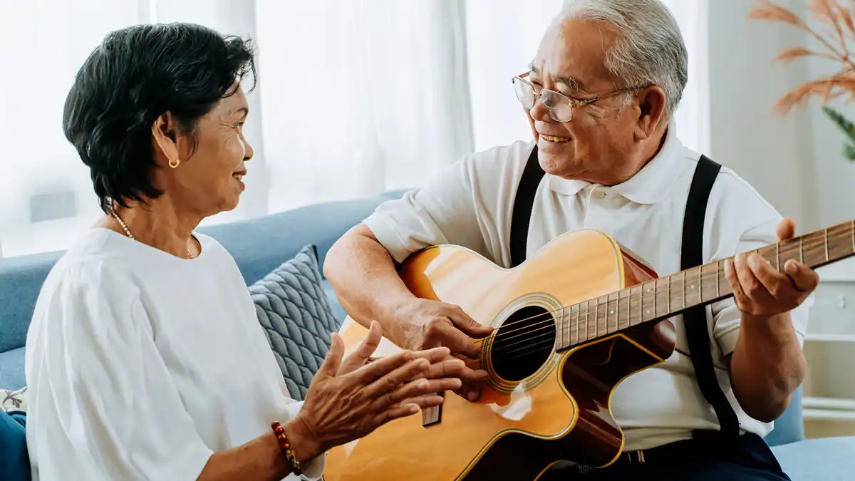 Two seniors sit together on a couch at home; one plays an acoustic guitar while the other listens and gestures, enjoying a relaxed musical moment.