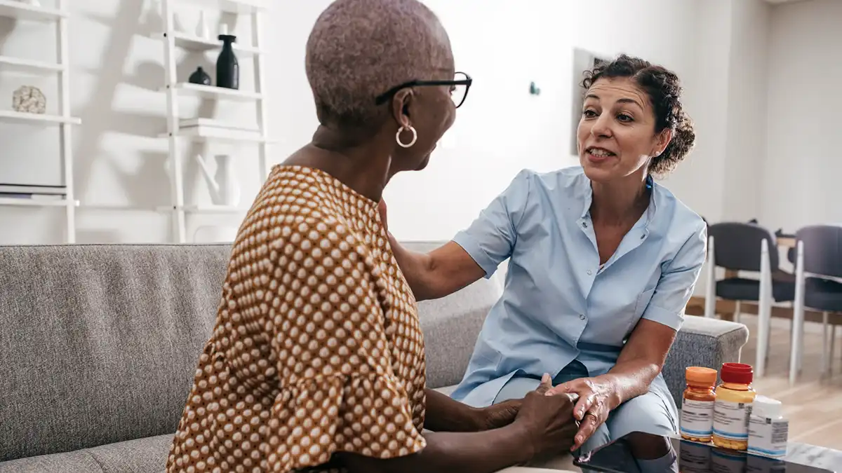 Compassionate nurse discusses medications with an older woman at home, offering supportive, collaborative care during a living room visit with pill bottles visible.