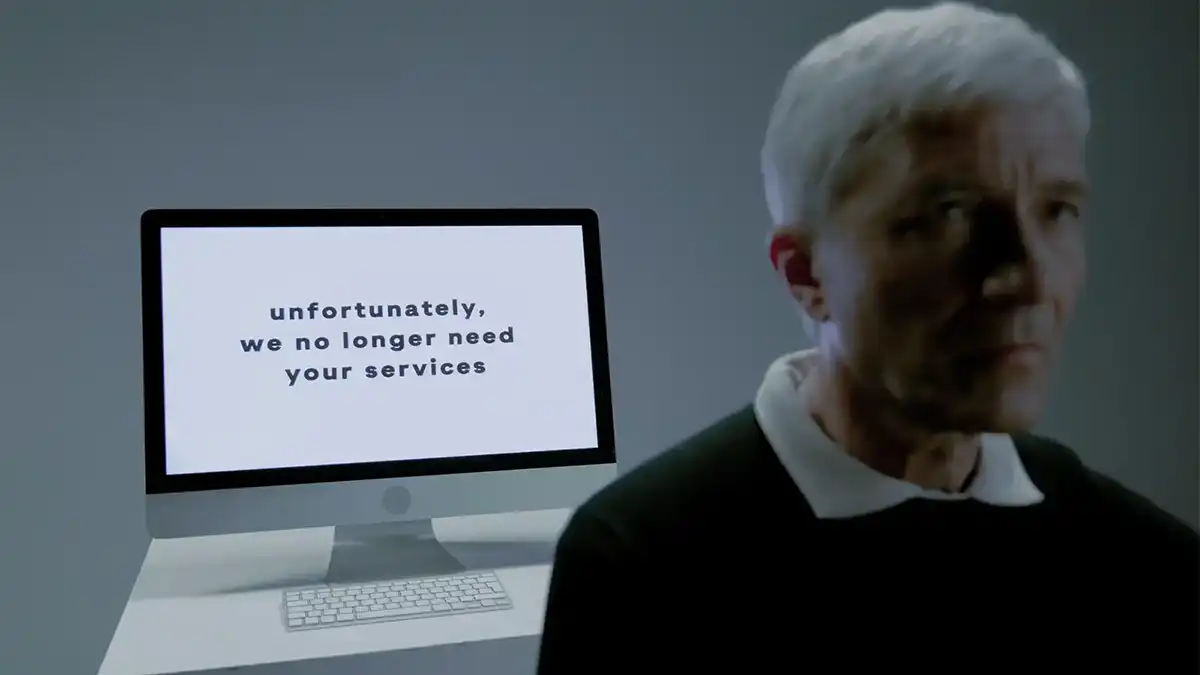 A somber man sits next to a monitor displaying the message: "unfortunately, we no longer need your services," representing a sudden job termination.