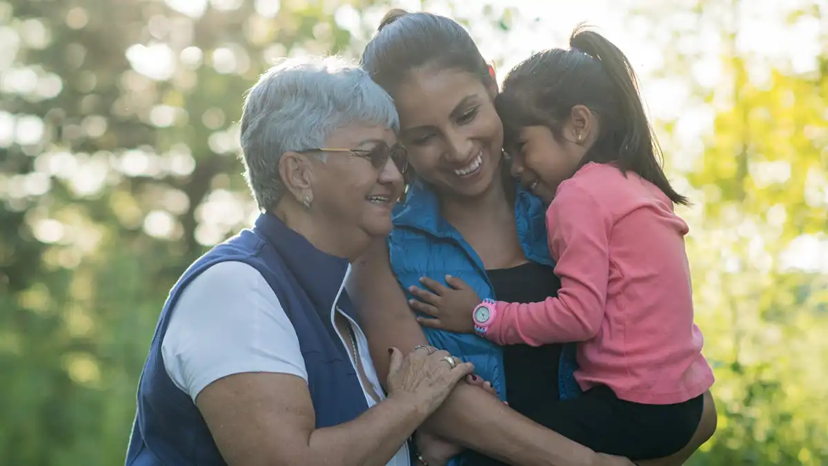 Smiling grandmother, mother, and young daughter embracing outdoors in a sunlit park, showcasing a loving three-generation family moment.