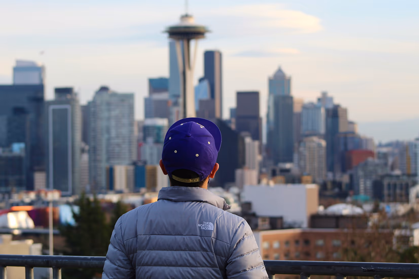 A photo of a young child wearing a purple baseball had facing away from the camera as he's looking at the Space Needle in the distance.
