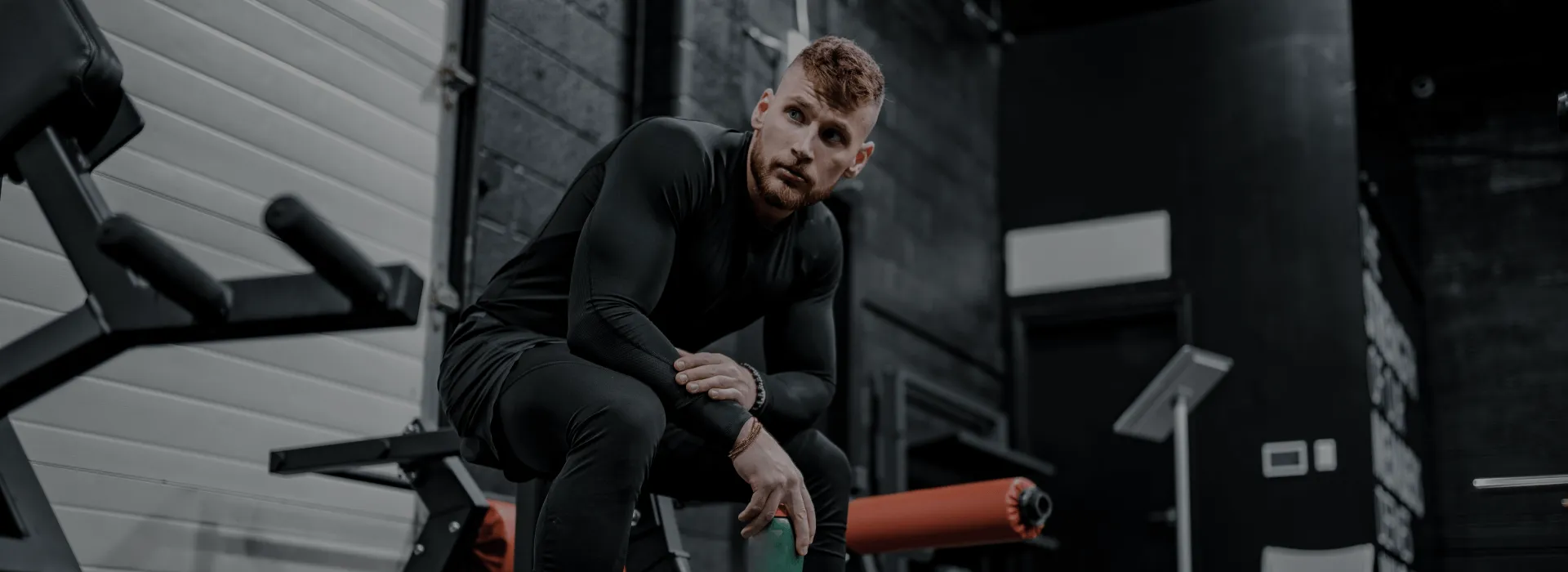 Man in black athletic wear sitting on a bench in a gym holding a green water bottle.