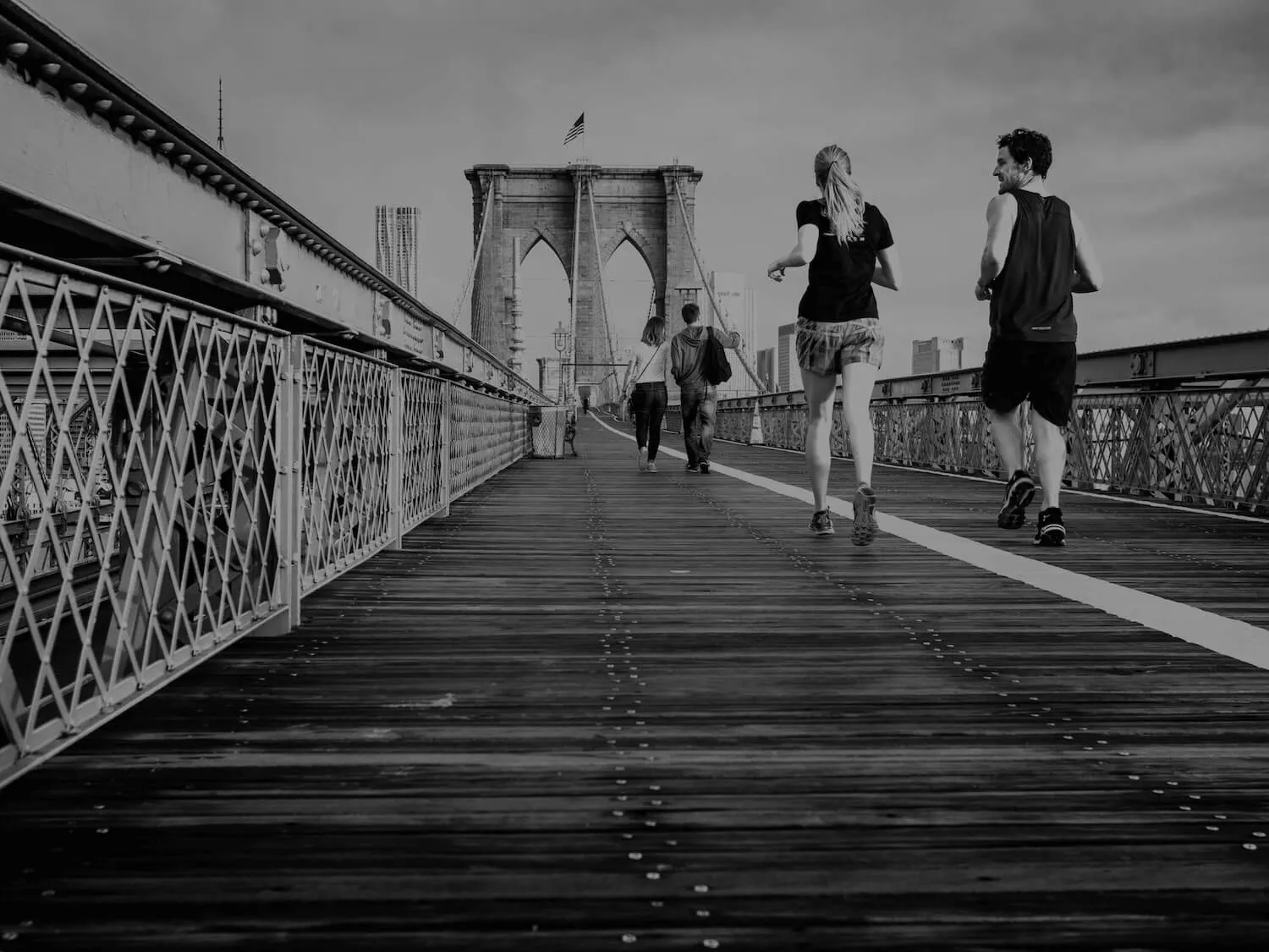 Two people jogging and two people walking on the wooden pedestrian path of the Brooklyn Bridge with city buildings in the background.