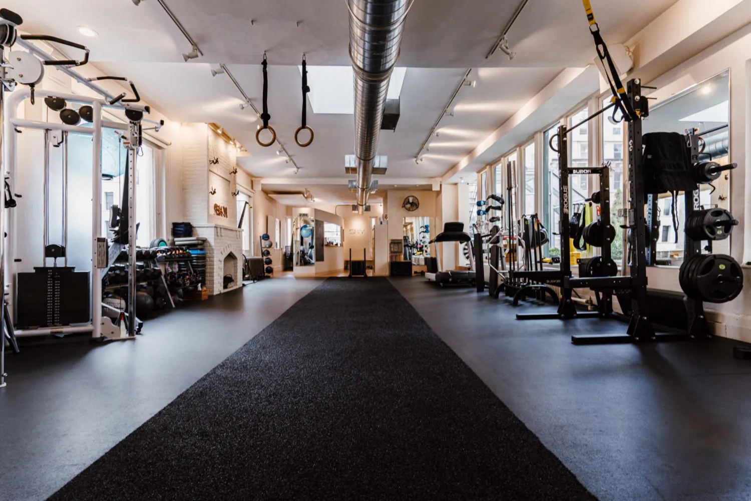 Modern gym interior with black rubber flooring, weight machines, dumbbells, kettlebells, gymnastic rings, and large windows along the right wall.