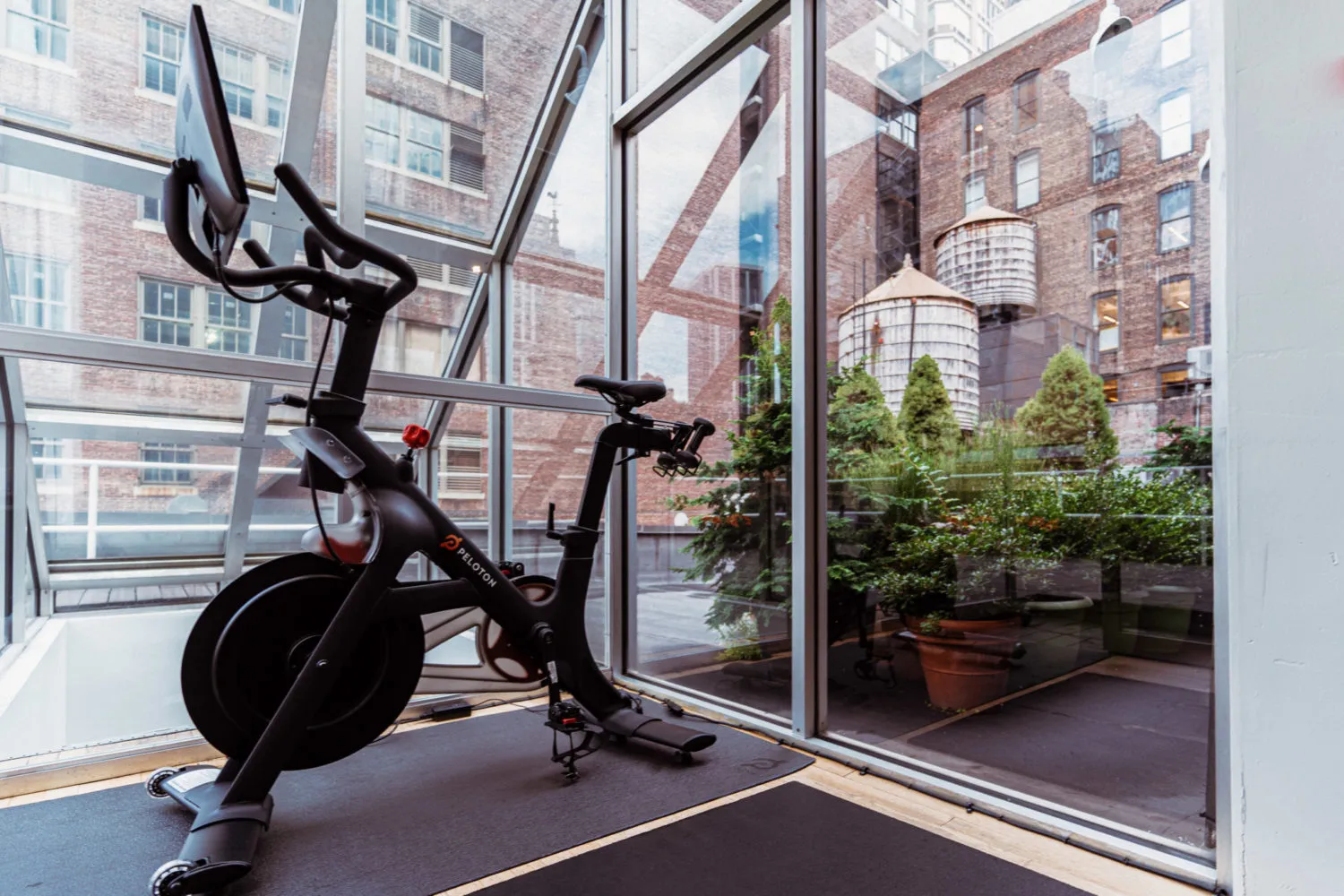 Black Peloton exercise bike inside a glass-walled room with an urban outdoor view of brick buildings and greenery.