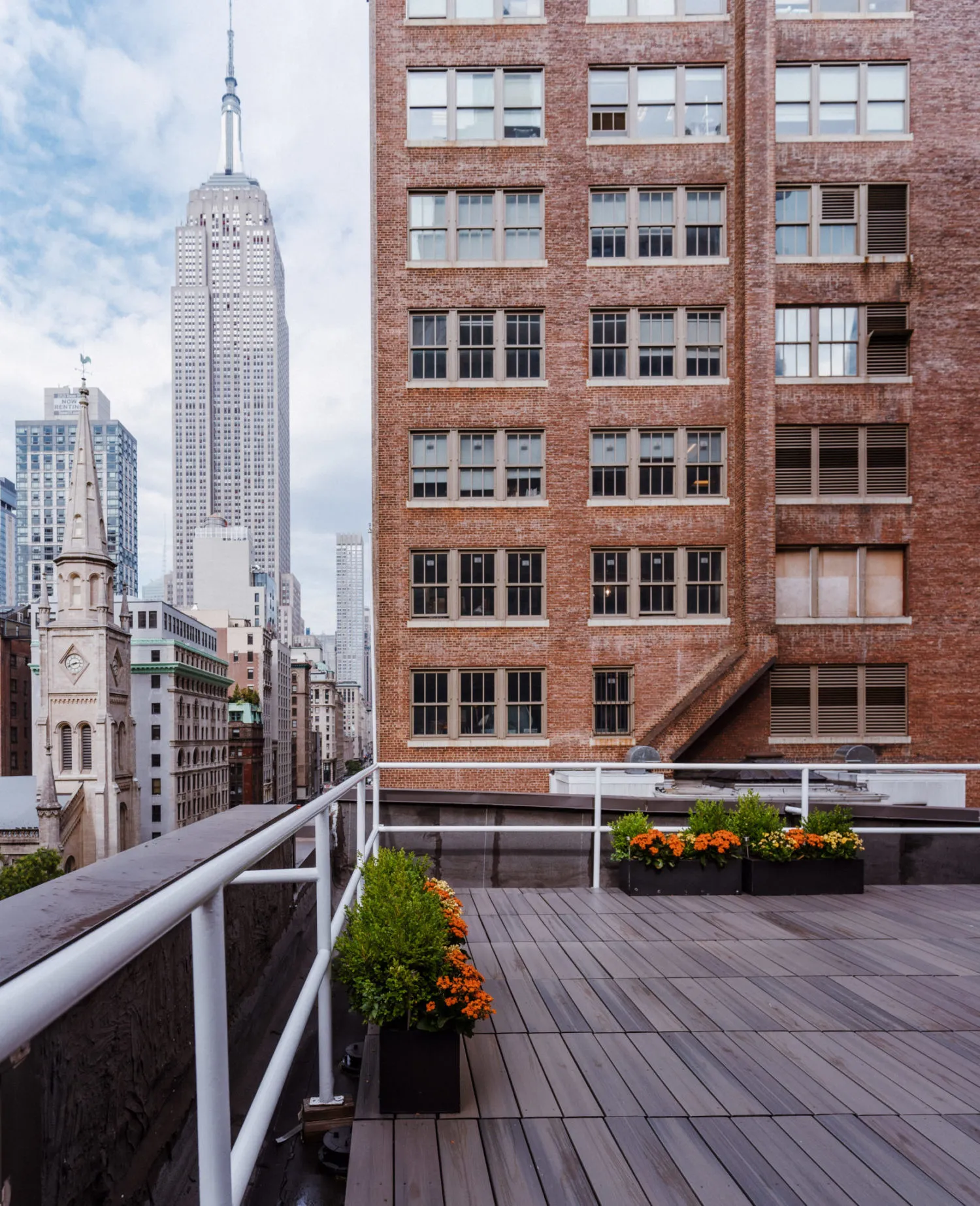 City rooftop terrace with wooden decking, rectangular planters of green bushes and orange flowers, and a view of the Empire State Building and surrounding buildings under a cloudy sky.