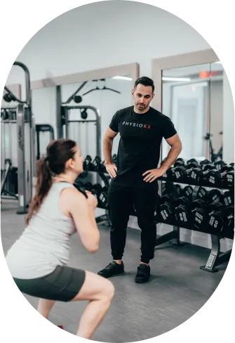 Male fitness trainer watches and coaches a woman performing a squat exercise in a gym with dumbbells and mirrors in the background.