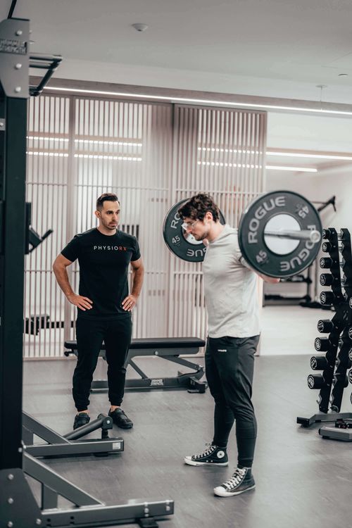Trainer watching a man perform a back squat with a loaded barbell in a gym.
