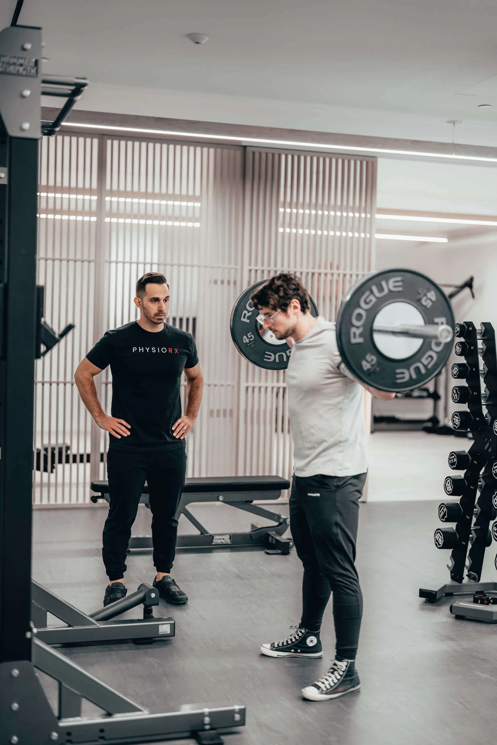 Trainer watching a man perform a back squat with a loaded barbell in a gym.