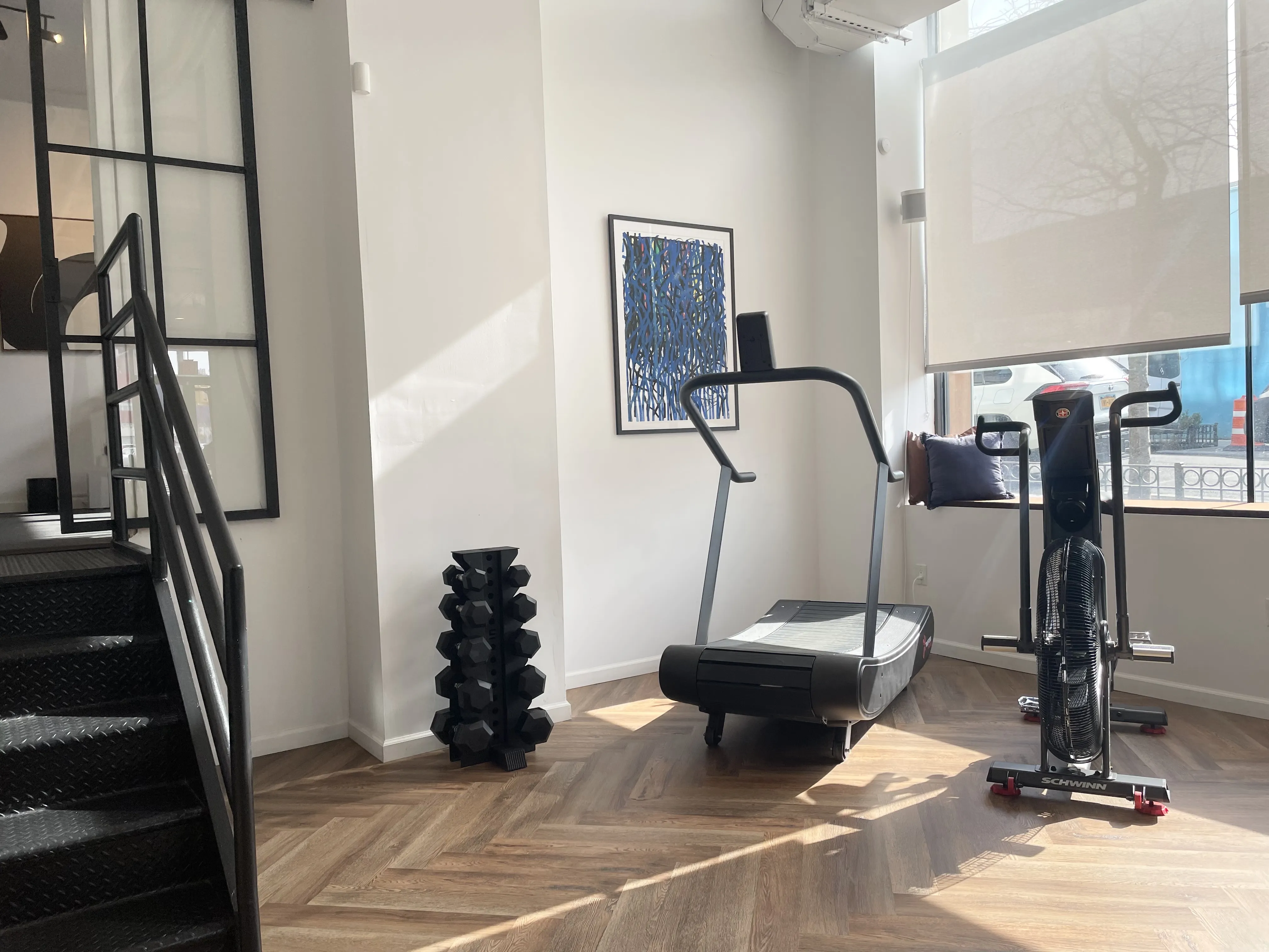 Bright gym corner with a curved treadmill, an air resistance exercise bike, and a rack of dumbbells next to a staircase.