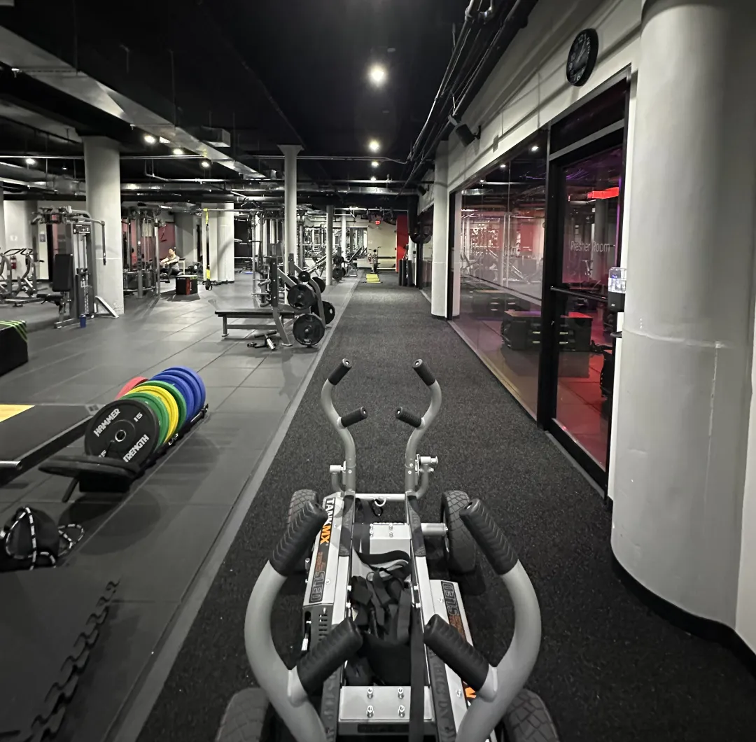Interior view of a spacious gym with weightlifting equipment, colorful weight plates, and a treadmill track alongside a glass-walled presser room.