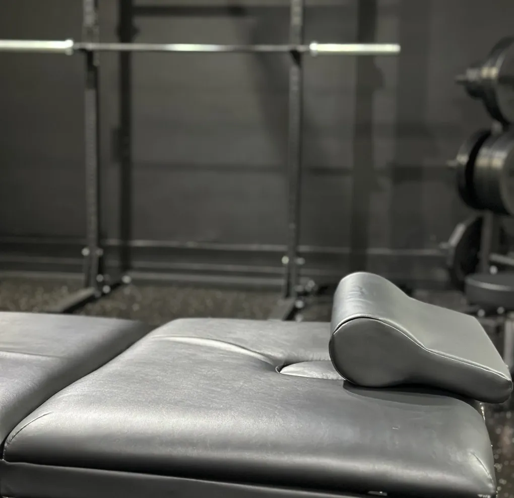 Close-up of a black padded gym bench with a small headrest, with weight rack and barbell blurred in the background.