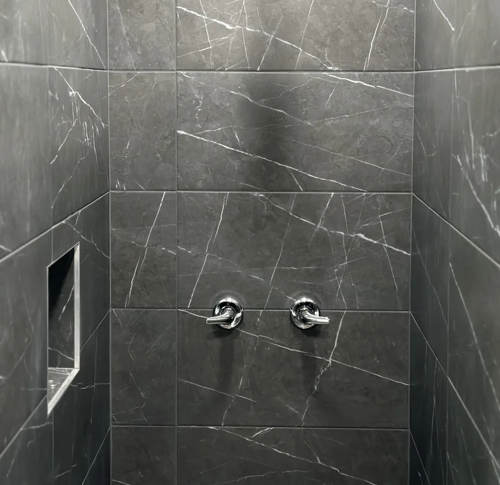 Dark gray marble tiled shower wall with two chrome water handles and a built-in recessed shelf on the left side.