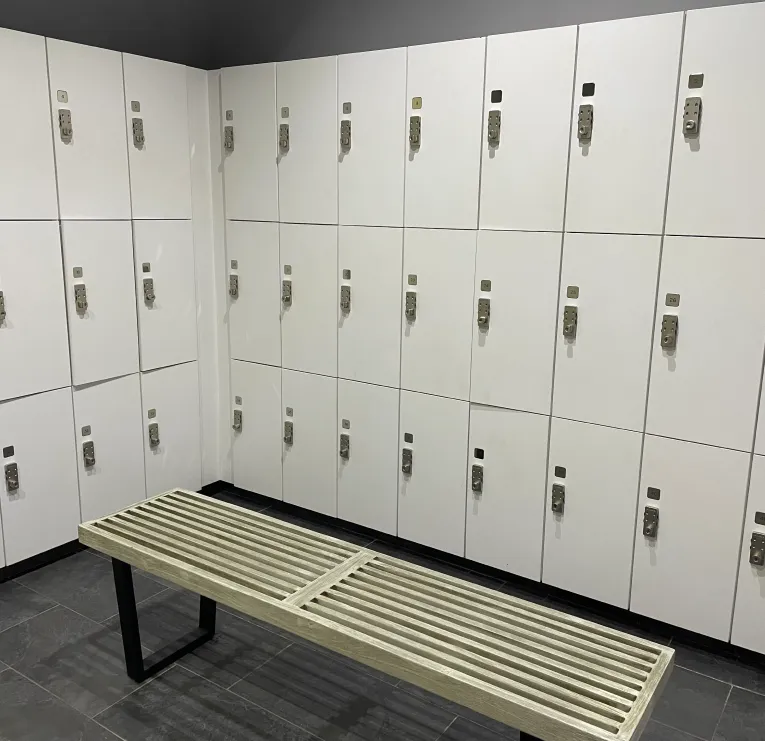 Corner of a locker room with white lockers and a wooden bench with black metal legs on a dark tiled floor.