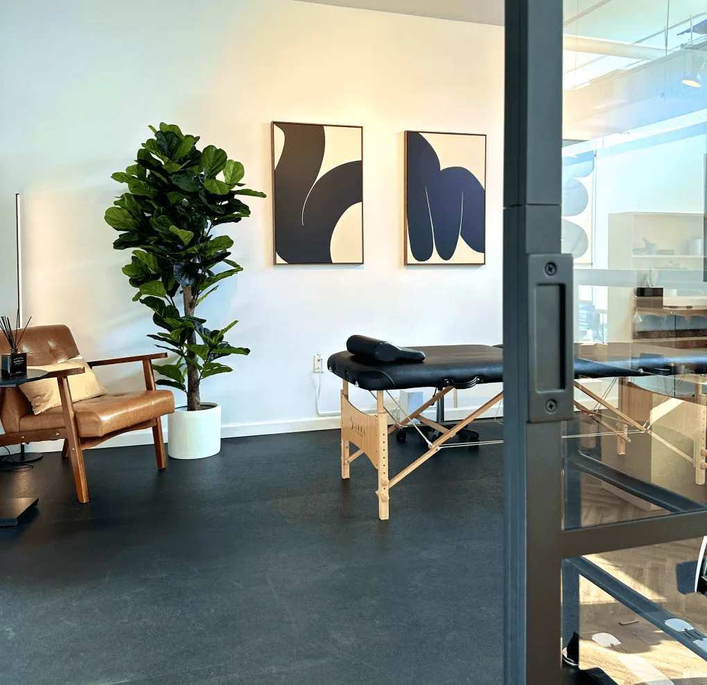 Minimalist therapy or massage room with a black massage table, wooden armchair with cushion, large potted plant, and two abstract black and beige paintings on the wall.