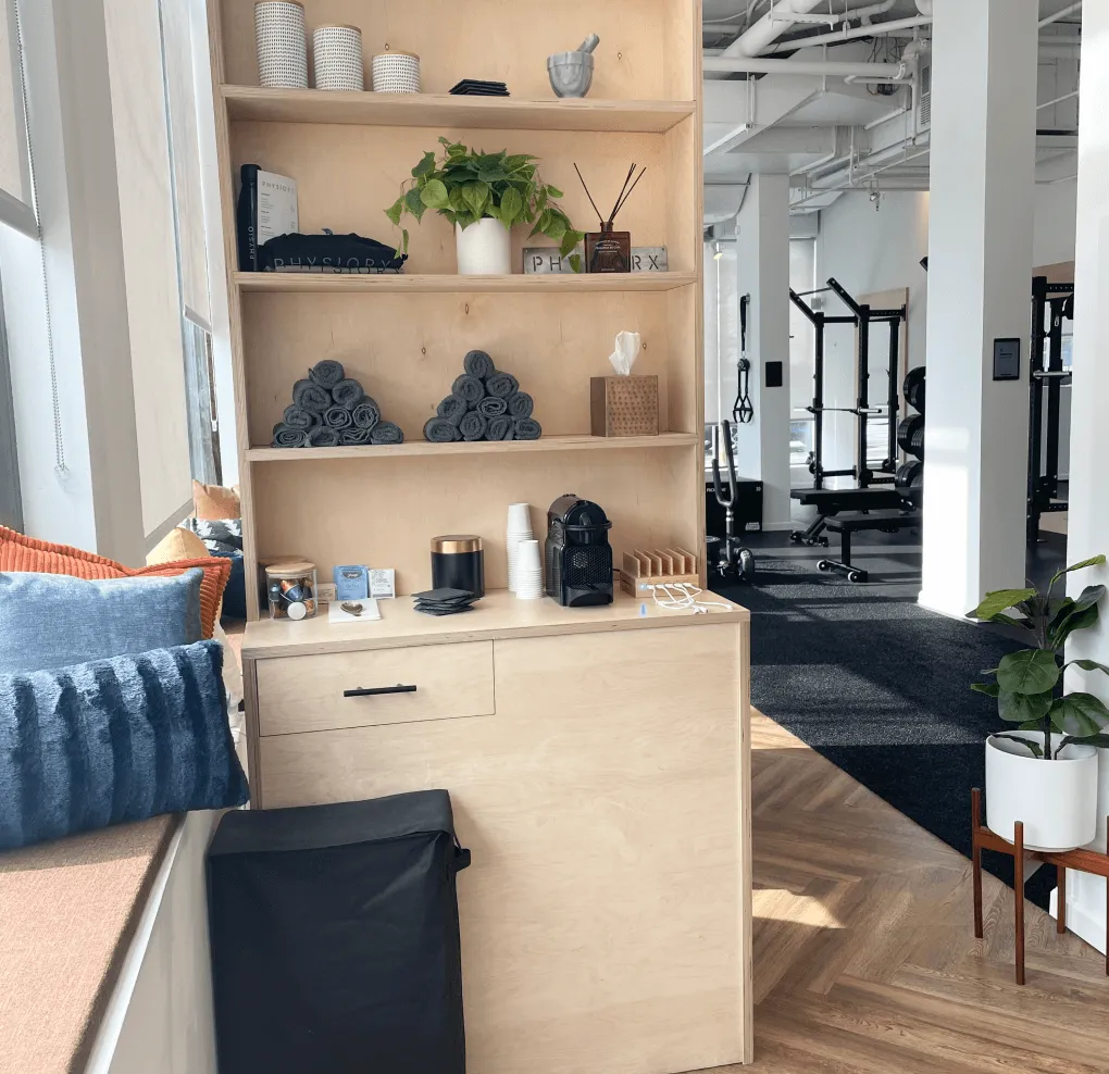 Light wooden shelves with rolled gray towels, a potted plant, cups, and coffee supplies in a bright gym area with weightlifting equipment and a plant on the right.