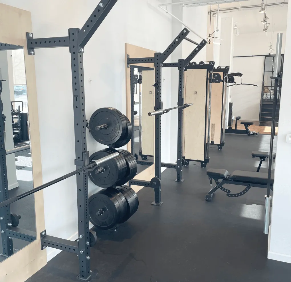 Indoor gym area with weightlifting racks, loaded barbells, benches, and exercise machines on black flooring.