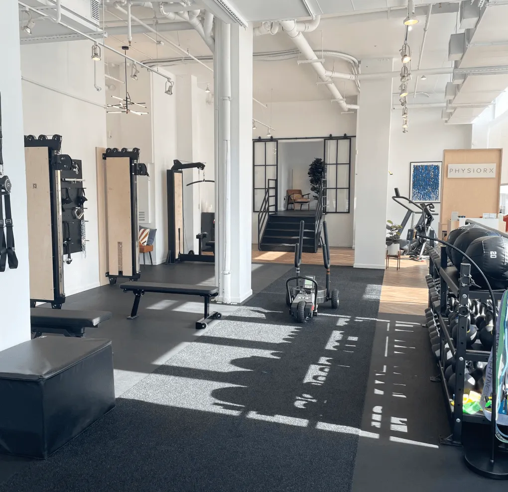 Bright gym interior with weight benches, exercise equipment, kettlebells, and a sign reading PHYSIORX on a light wood partition.