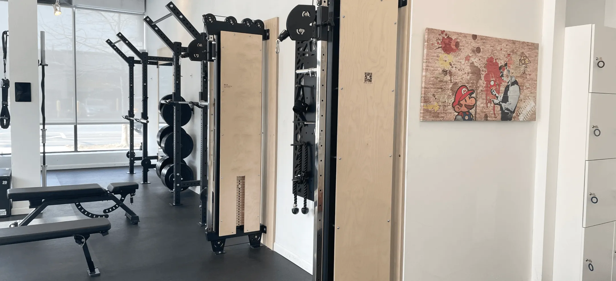 Modern gym interior with weight benches, squat racks holding weight plates, pulley machines, and a wall artwork featuring Mario from Nintendo.