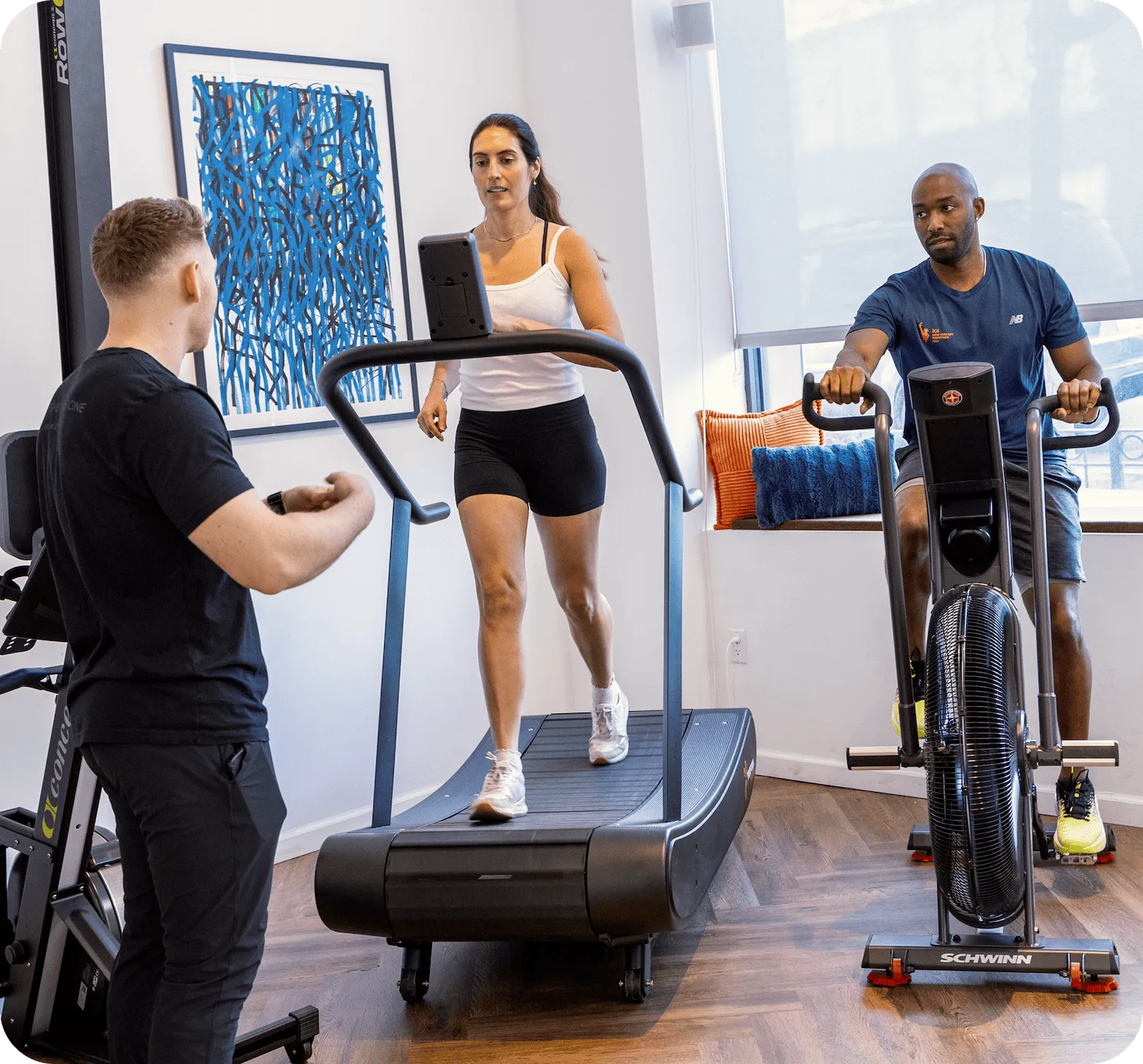 Coach guiding a woman on a treadmill while a man rides an air bike in a bright studio