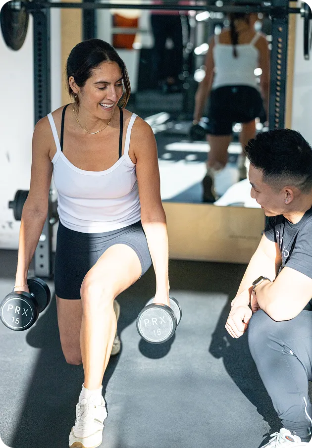 Woman doing a dumbbell lunge while a coach kneels beside her giving guidance