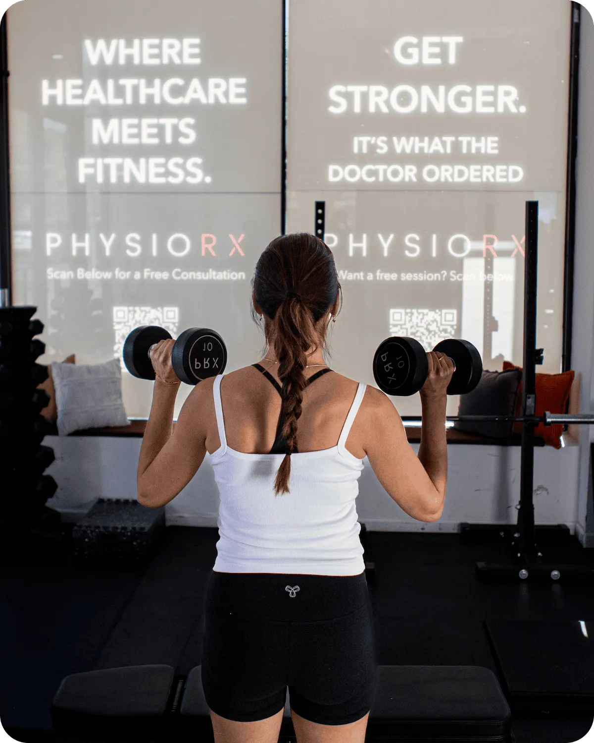 Woman doing a dumbbell shoulder press facing PhysioRX window signs: Where Healthcare Meets Fitness