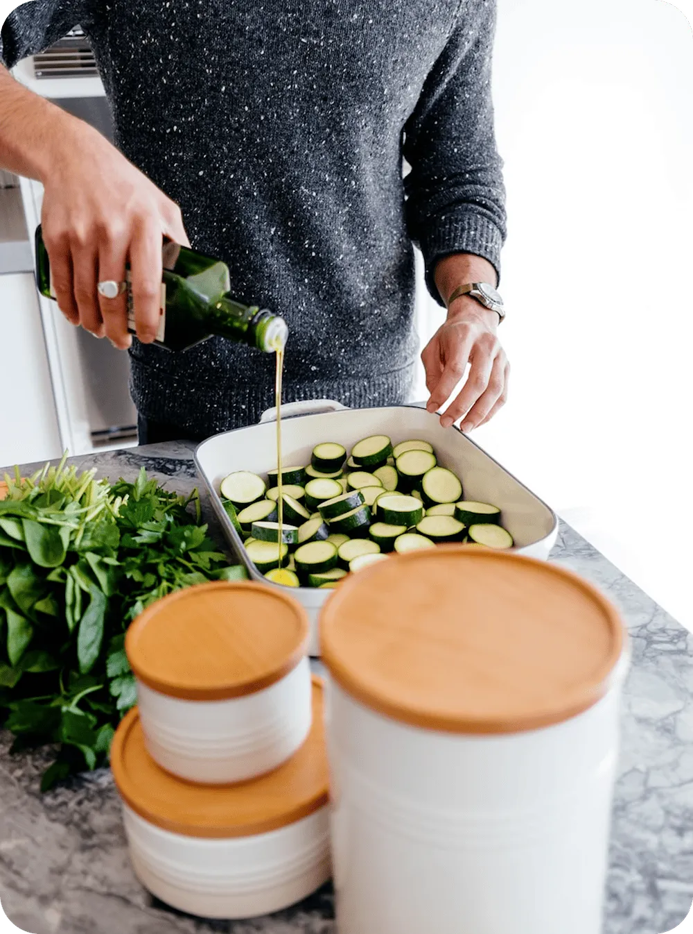 Hands drizzling olive oil over sliced zucchini in a baking dish