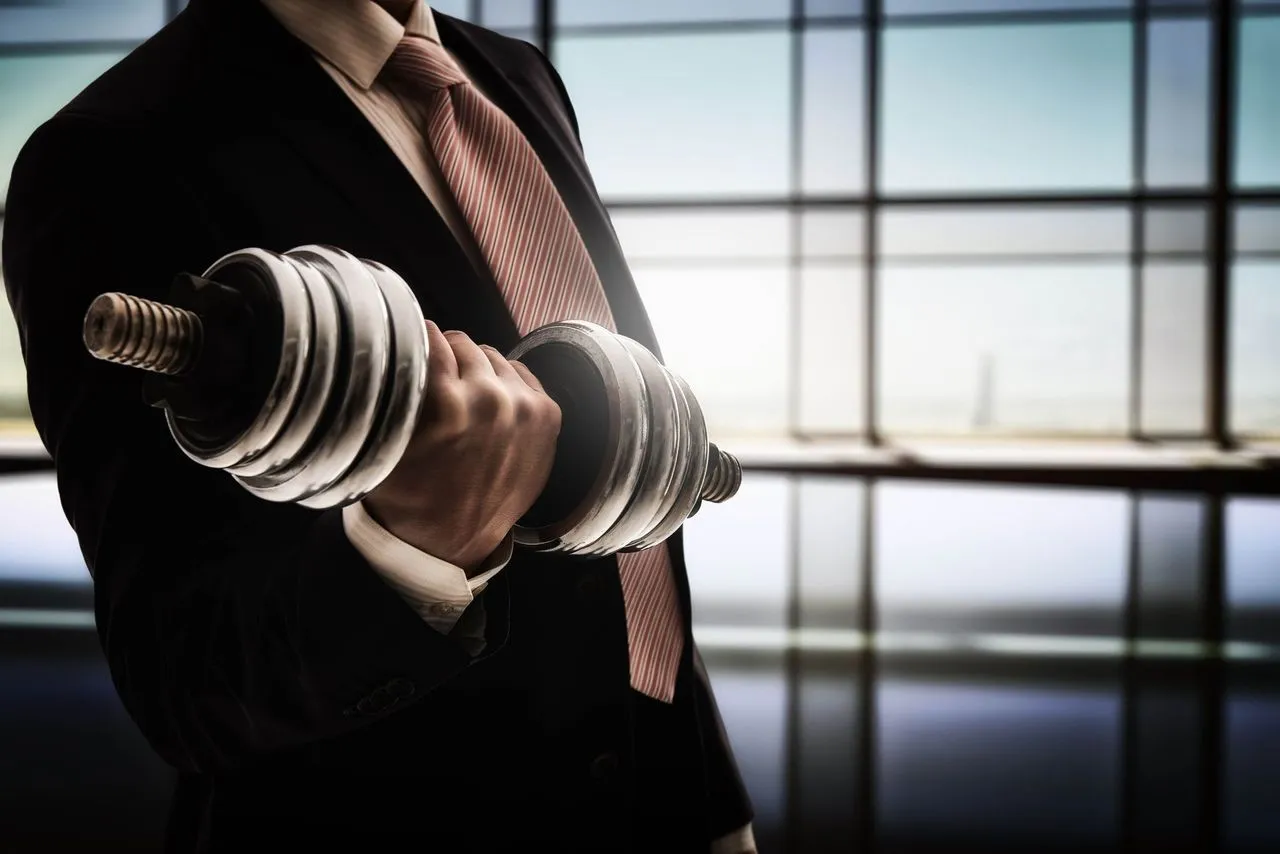 Businessman in a suit and tie lifting a dumbbell in front of large office windows