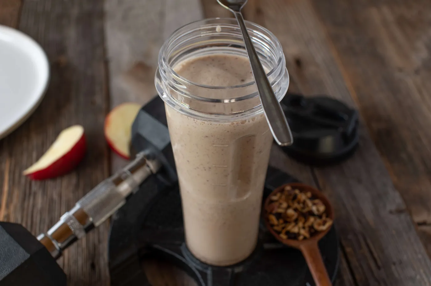 Protein shake in a blender cup next to apple slices, chopped nuts and a dumbbell on a wooden table