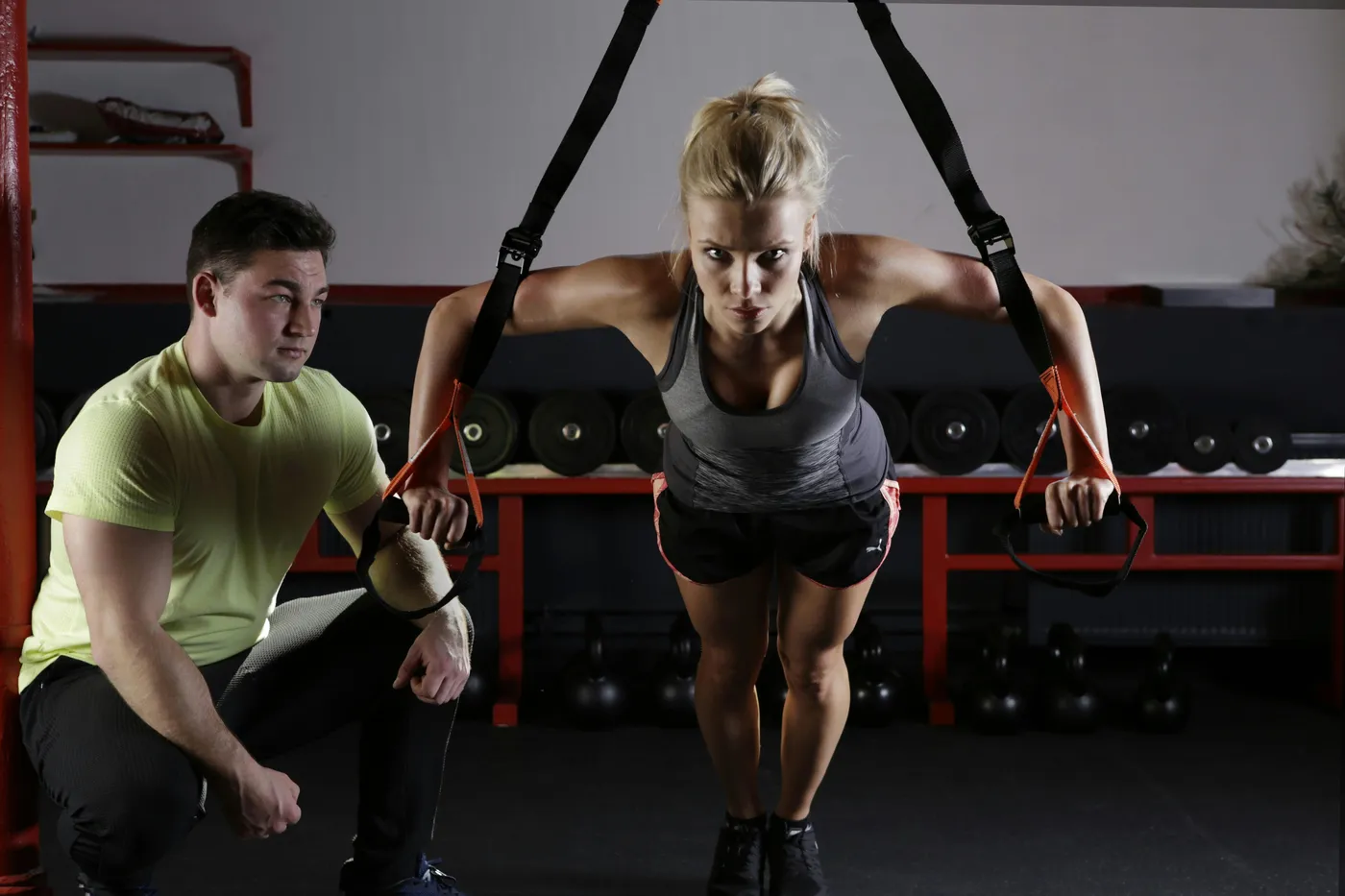 Trainer kneeling beside a woman doing a suspension strap chest press in a gym