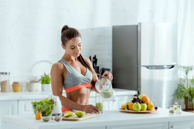 Fit woman pouring a green smoothie in a kitchen with fruit and vegetables on the counter