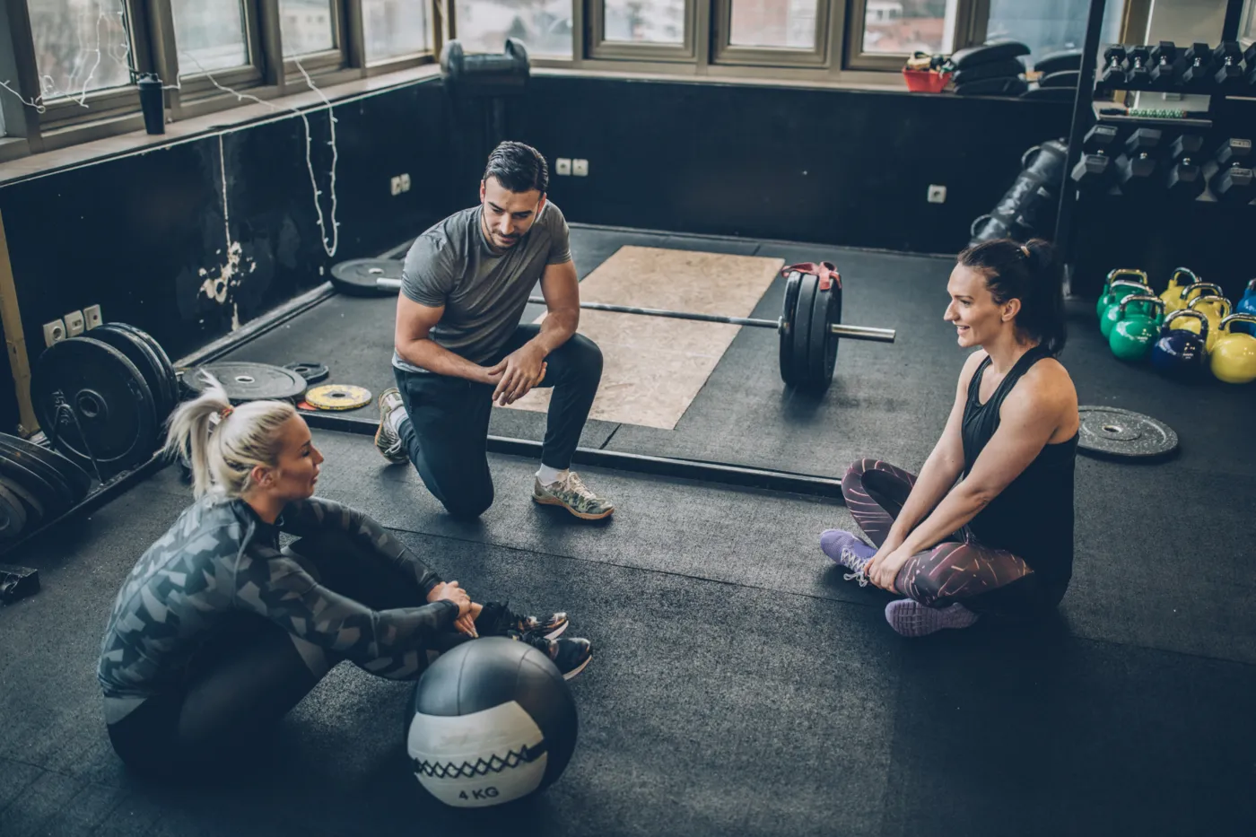 Coach kneeling to talk with two women seated on the floor of a gym with barbells and kettlebells