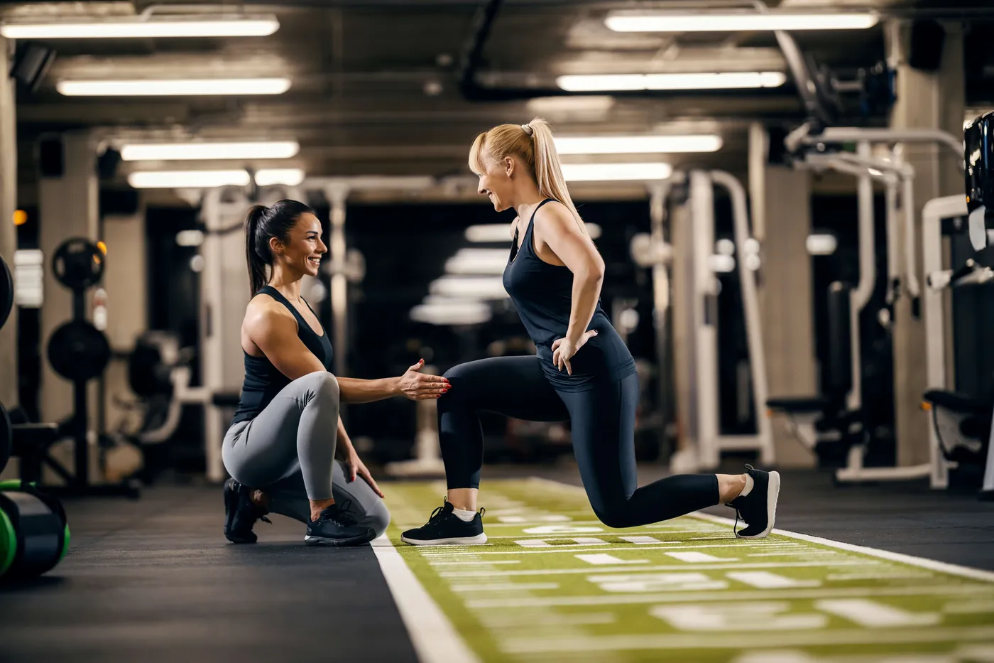 Woman guiding another woman through a lunge on a turf strip in a large gym