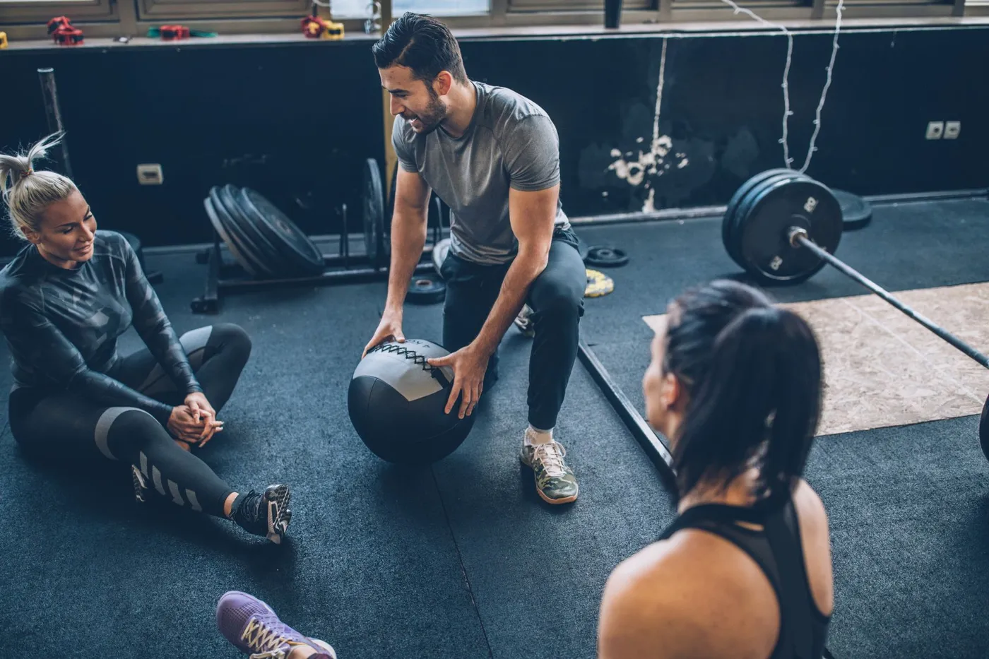 Coach holding a medicine ball while talking with two women sitting on a gym floor