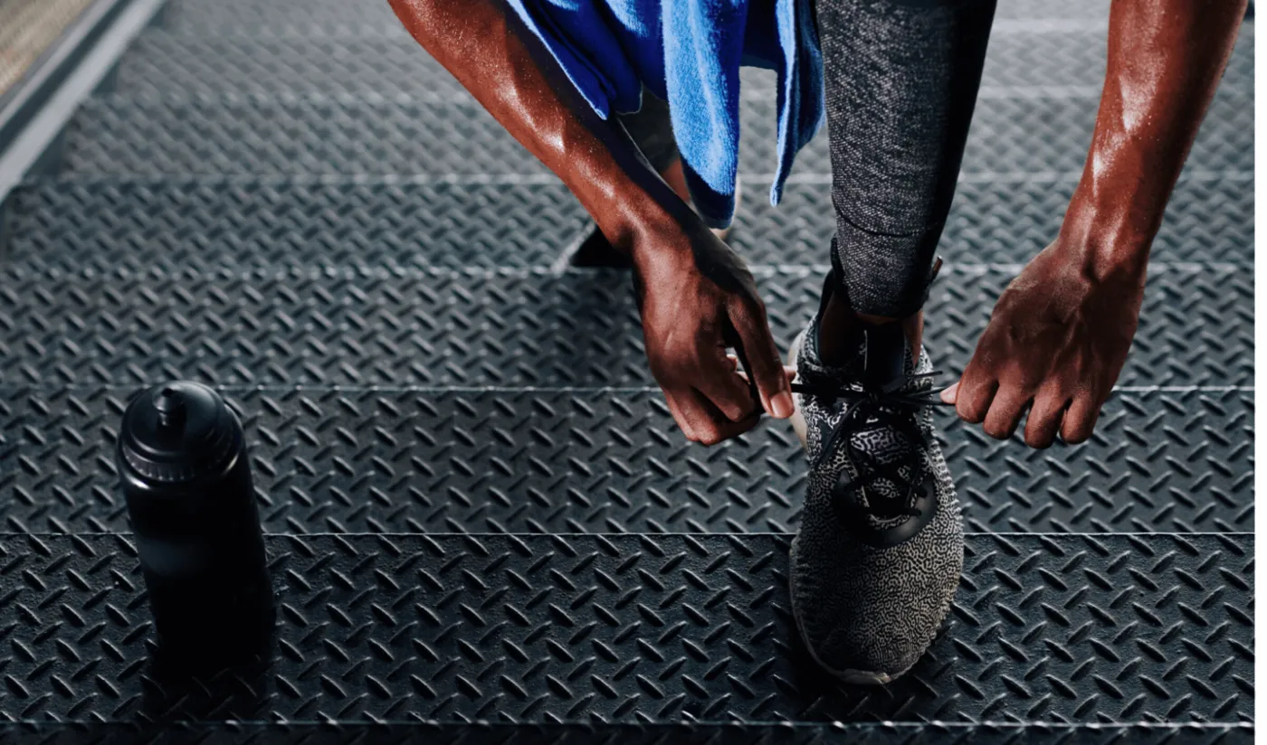 Athlete tying a running shoe on a metal treadmill-style floor beside a water bottle