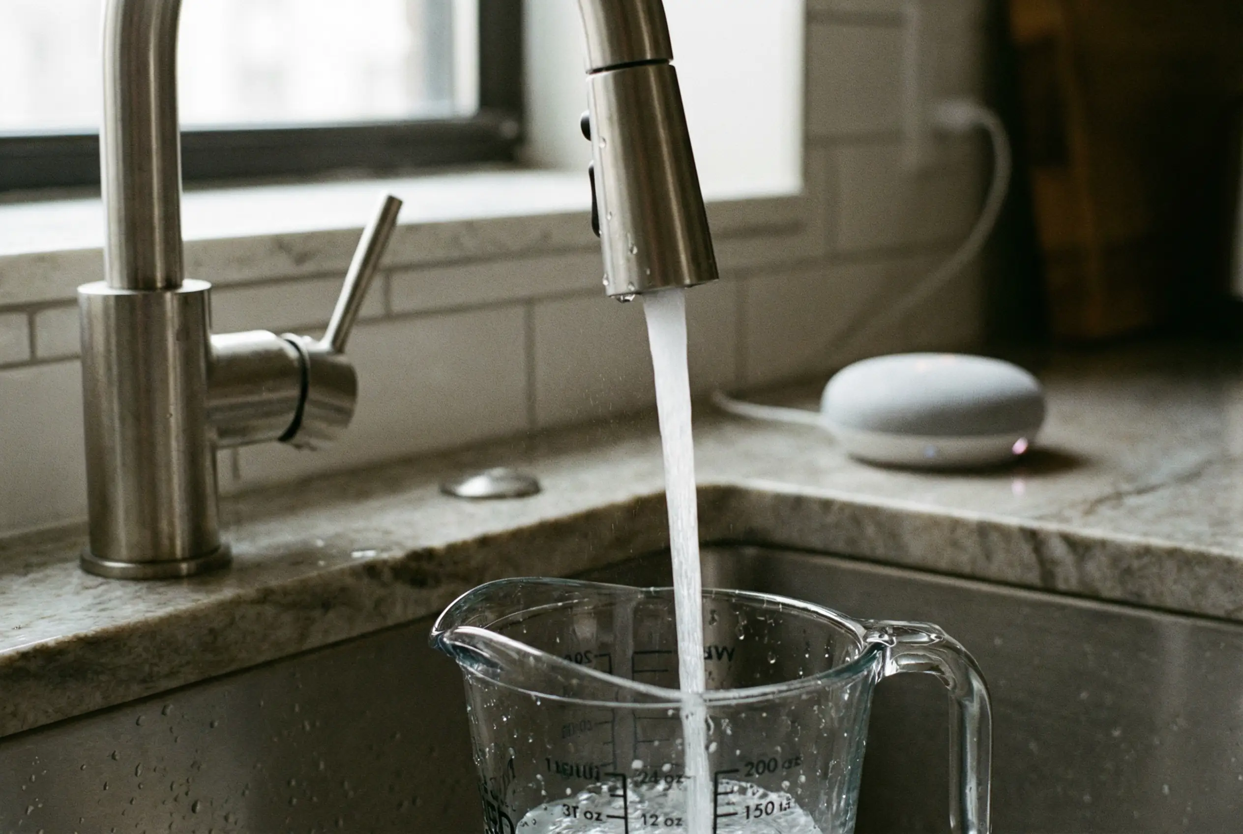 Faucet dispensing water into a measuring cup with a voice assistant nearby in a Manhattan kitchen.
