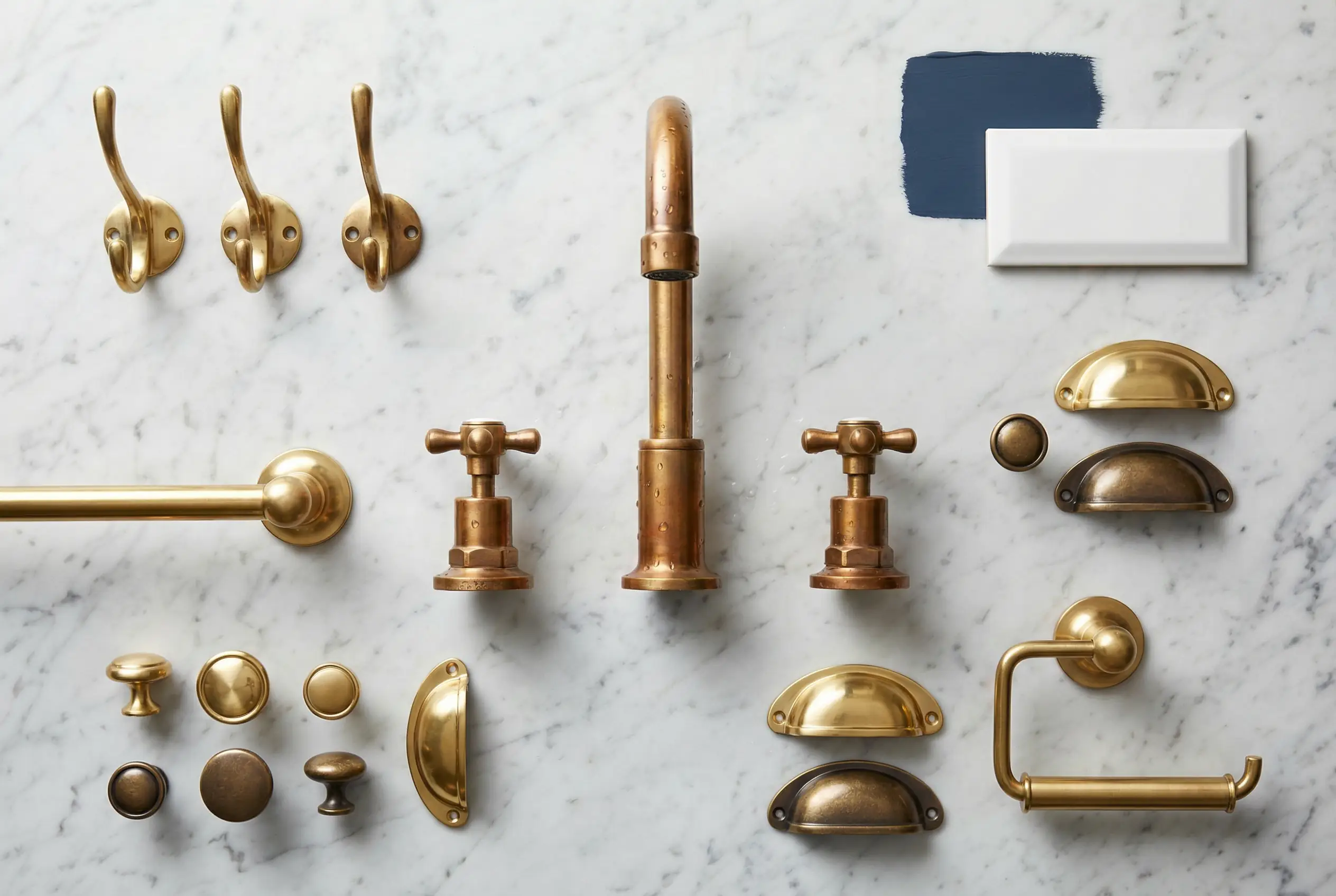 Flat lay of unlacquered brass bathroom fixtures including faucet, towel bar, and cabinet hardware on marble surface showing natural patina