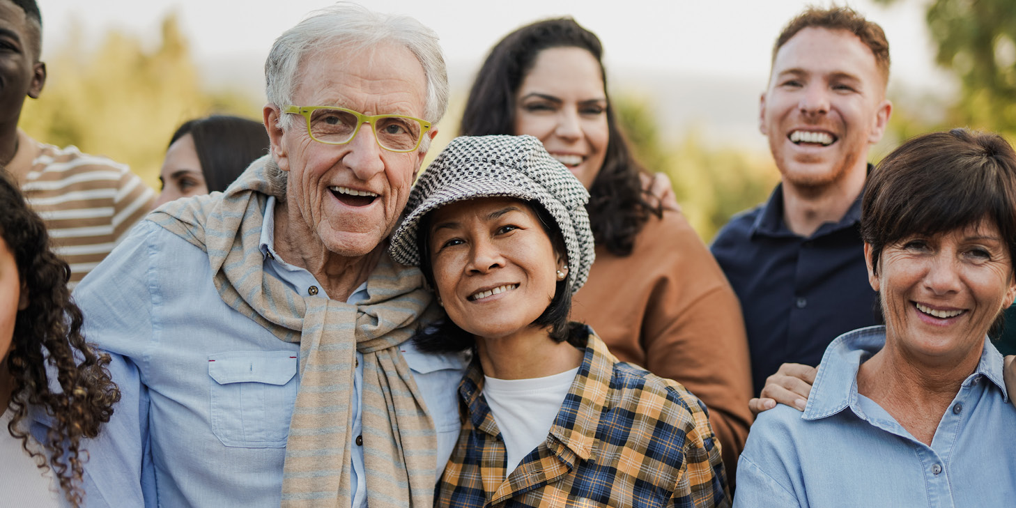 A diverse group of smiling people standing close together outdoors, including an elderly man with glasses, a woman in a plaid shirt and hat, and others in casual clothing.