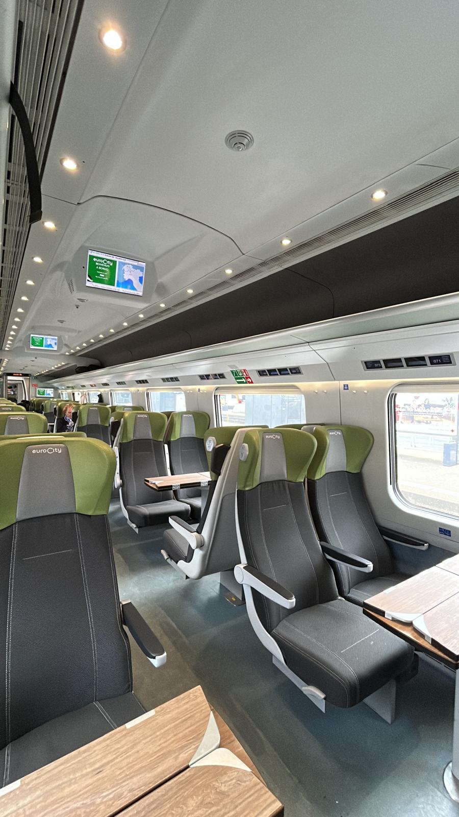 Modern Swiss EuroCity train interior with green-and-grey seats, tables and overhead information screens.
