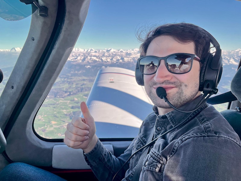 Giacomo Iotti gives a thumbs-up from a small aircraft cockpit, wearing a headset with snow-covered mountains behind him.