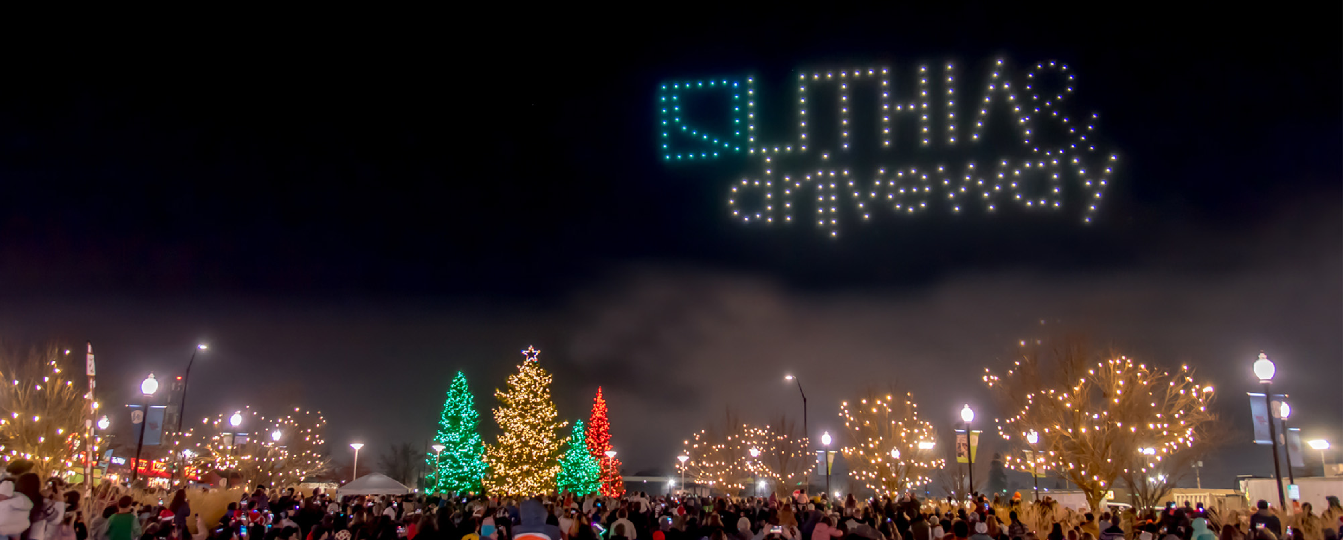 Image of Pear Blossom Park with trees lit up for the holidays under a sky with a drone light show of Lithia & Driveway's logo 