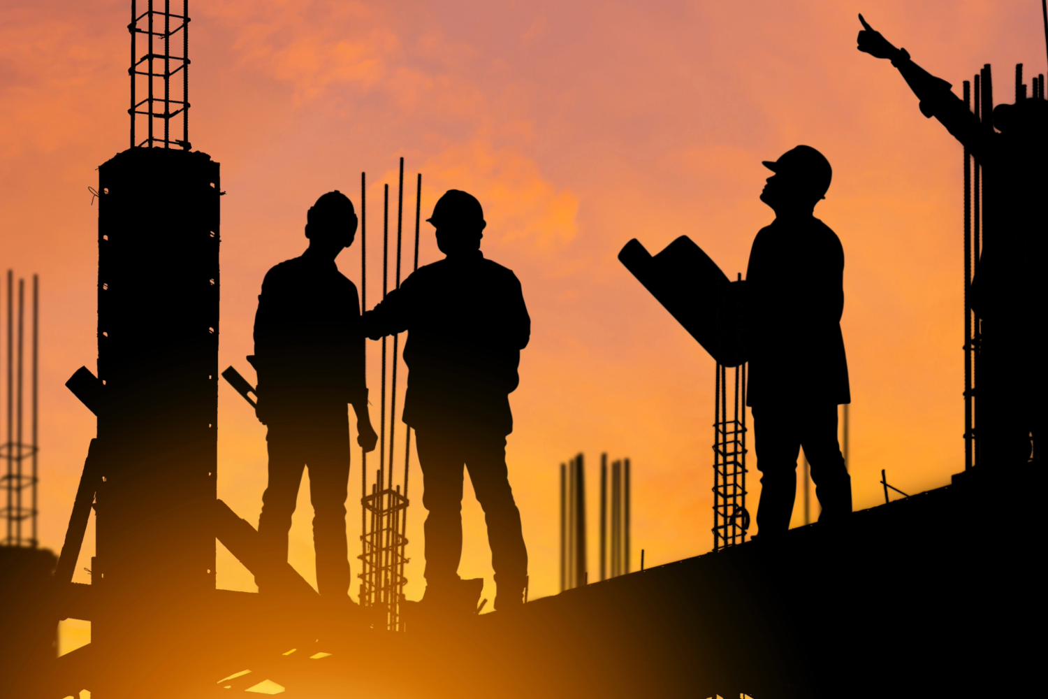 Construction workers silhouetted against a sunset, discussing plans on a construction site.