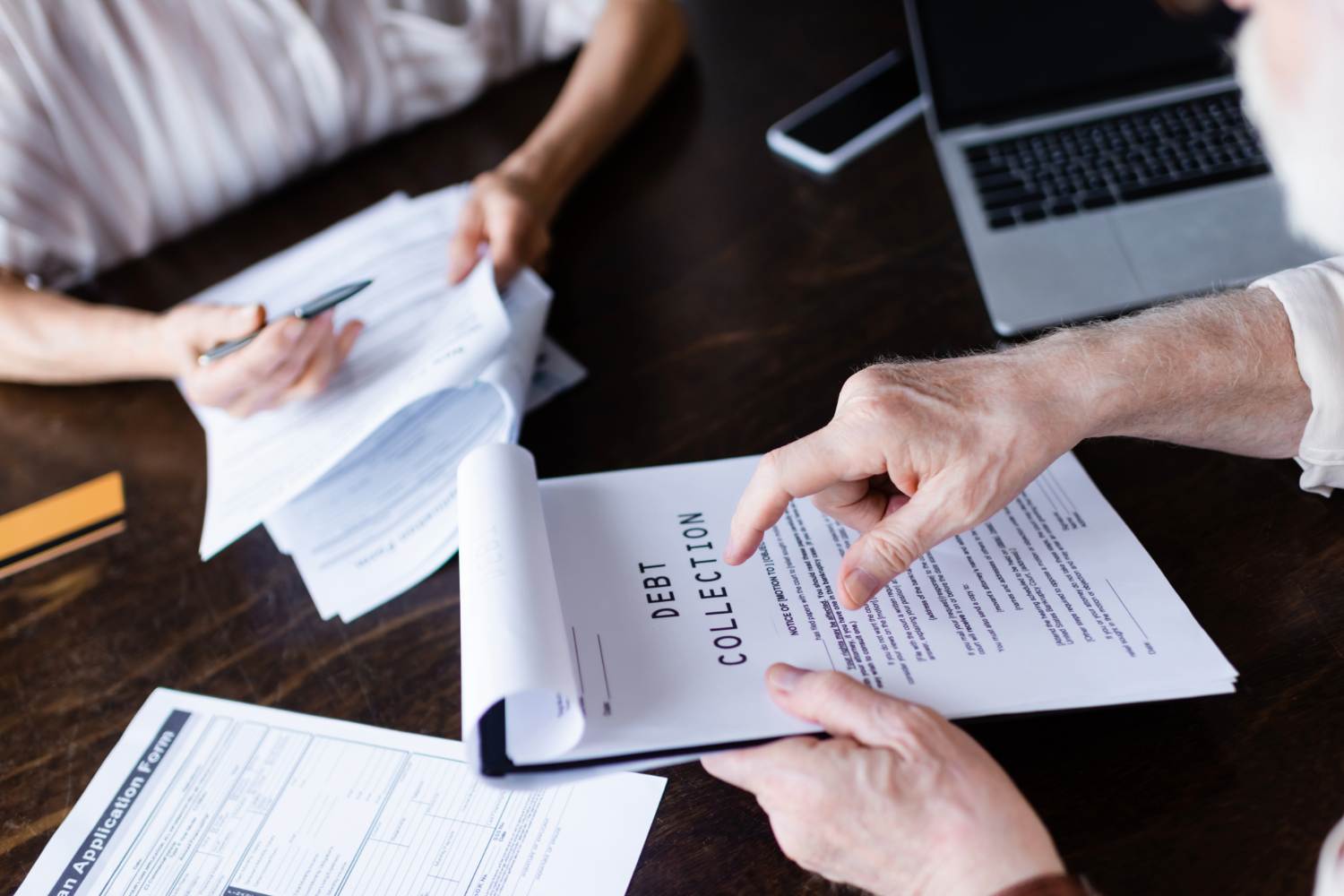 Hands pointing at "Debt Collection" document on a table with paperwork and a laptop.