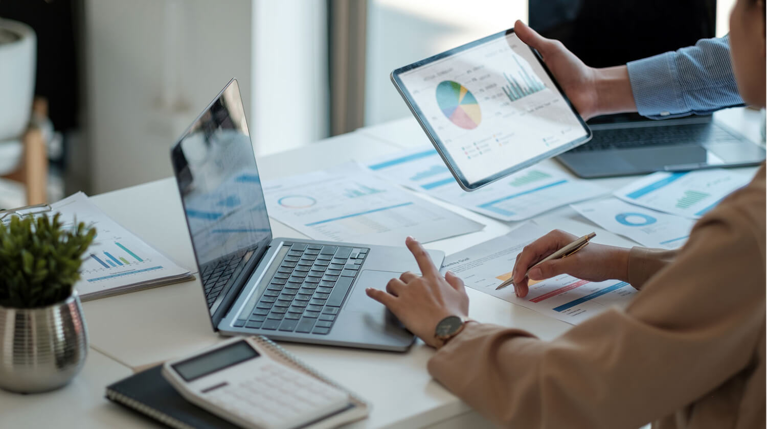 Person analyzing financial data on a laptop and tablet, surrounded by documents and charts.