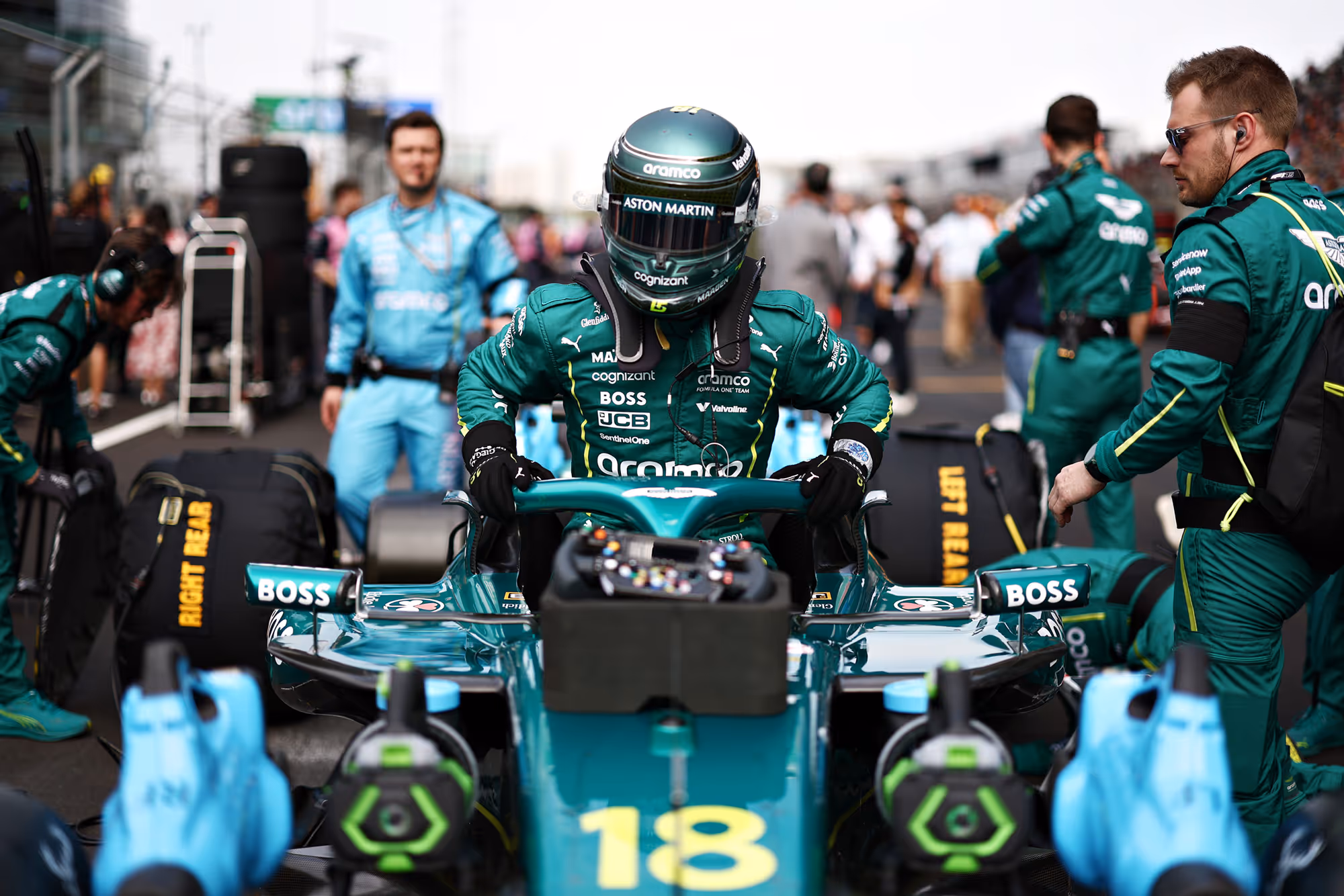 Aston Martin Aramco F1 driver sits in the teal race car on the grid surrounded by the pit crew and team members before the race.