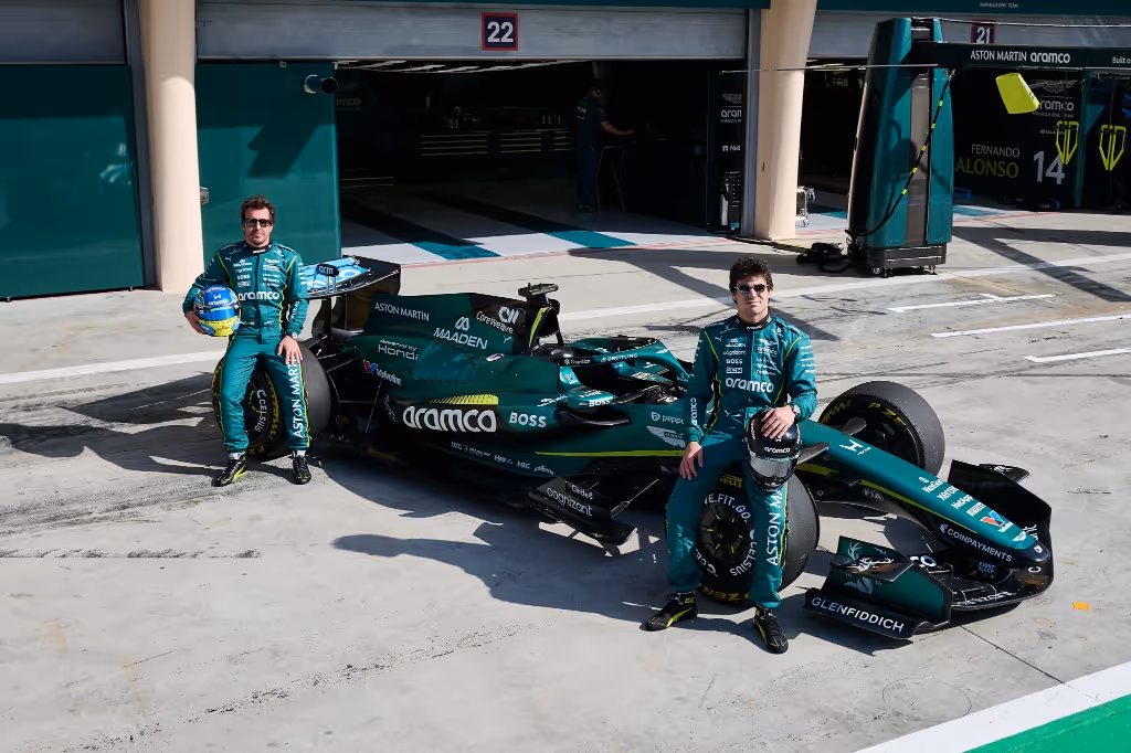 Two Formula 1 drivers in green racing suits posing with their green F1 car in front of a garage.
