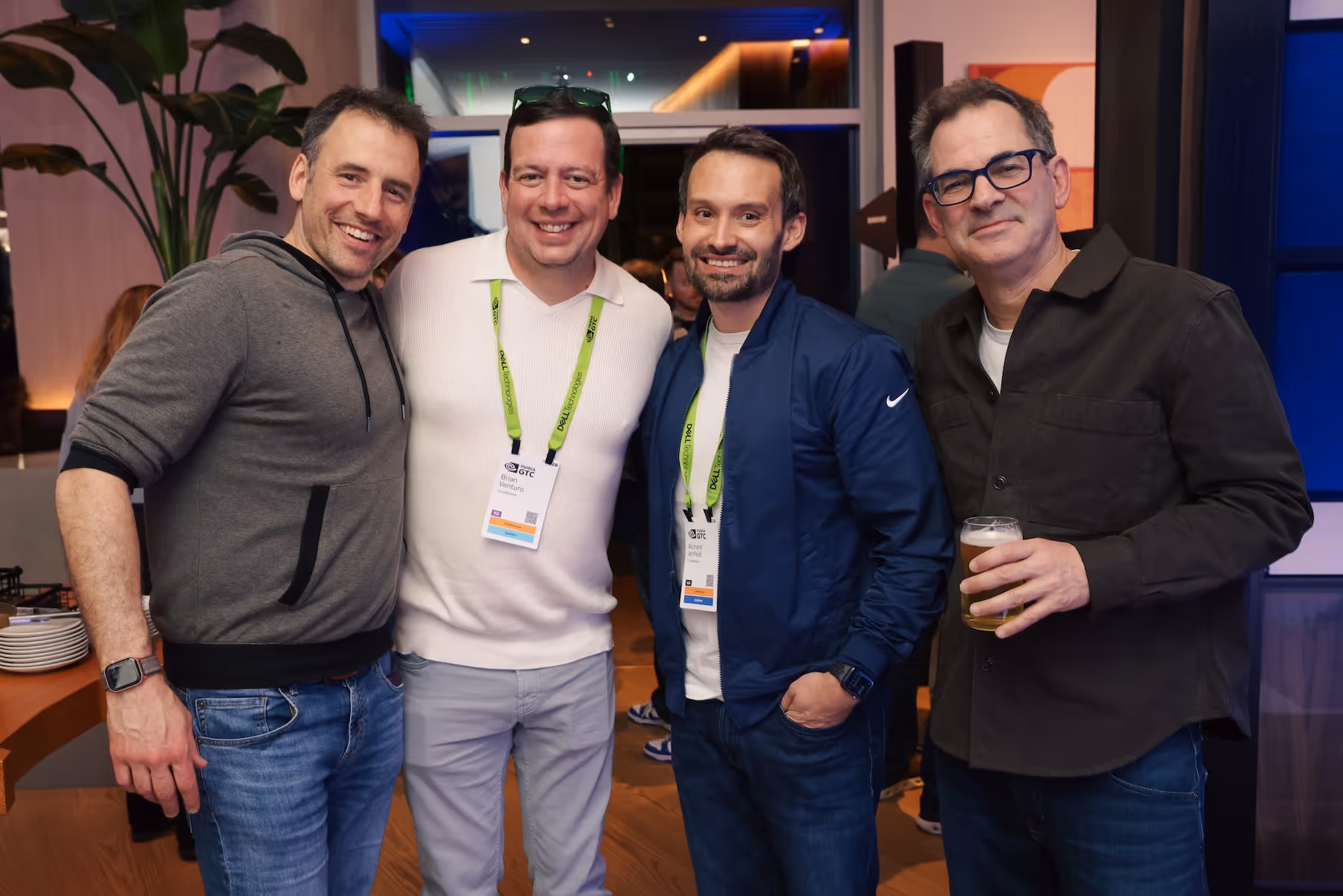Four men smiling and posing together at an indoor event, two wearing conference badges and one holding a glass of beer.